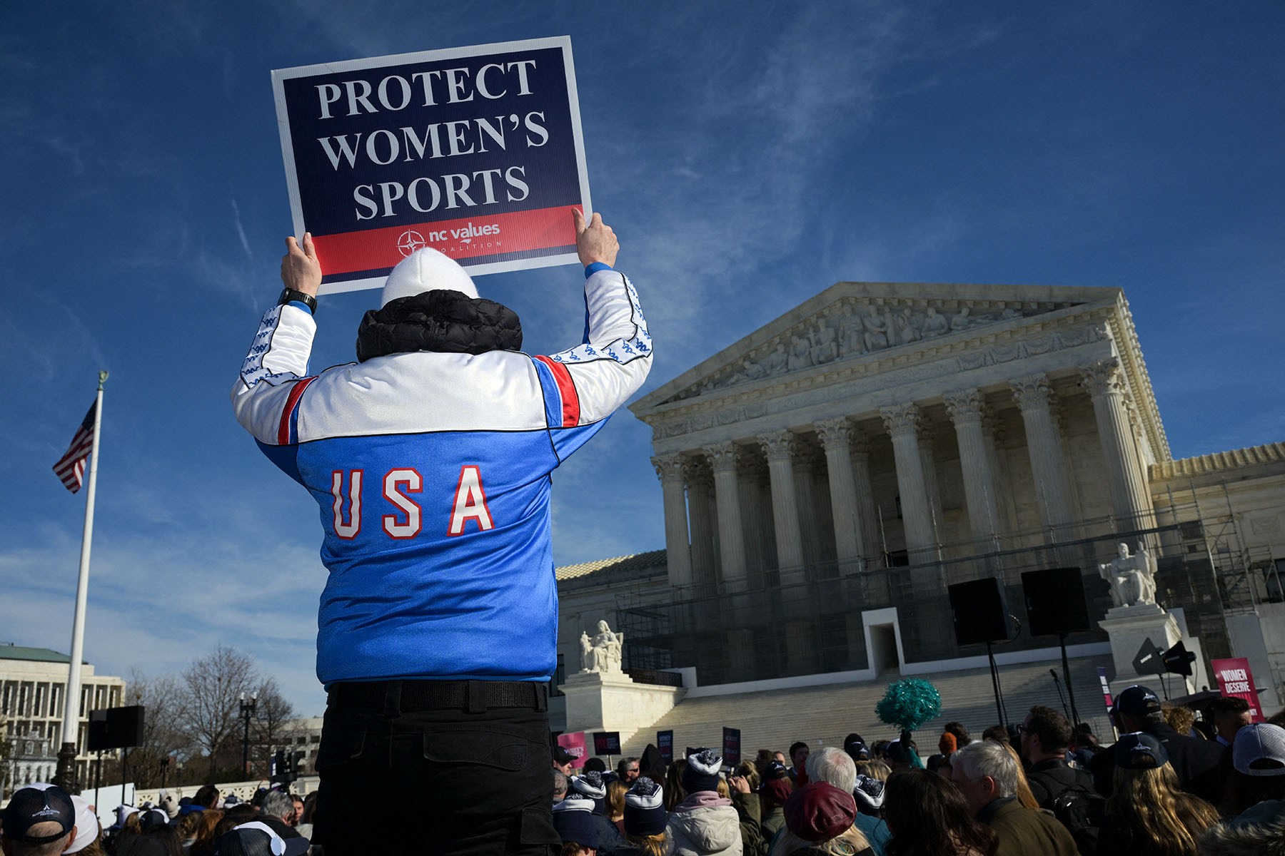 A person wearing a red, white and blue USA jacket holds a "Protect Women's Sports" sign outside the Supreme Court during a rally.