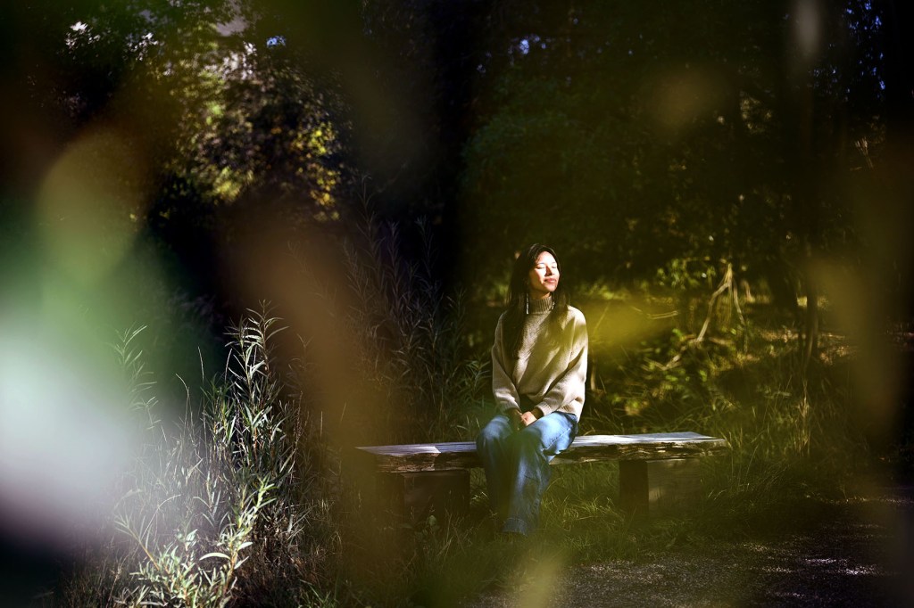 Katie Fire Thunder sits alone on a wooden bench in a wooded clearing, wearing a gray sweater and jeans. She is illuminated by a patch of sunlight, while out-of-focus foliage frames the scene in the foreground.