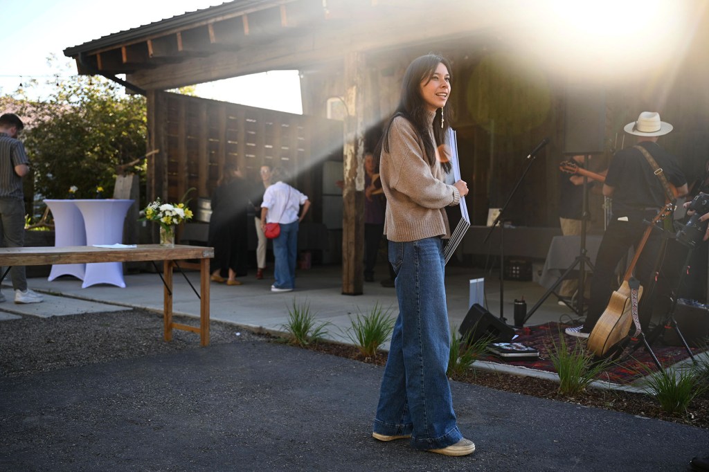 Katie Fire Thunder smiles while standing outside a rustic wooden building, holding campaign signs. Sunlight streams across the scene, with musical instruments and other attendees nearby.