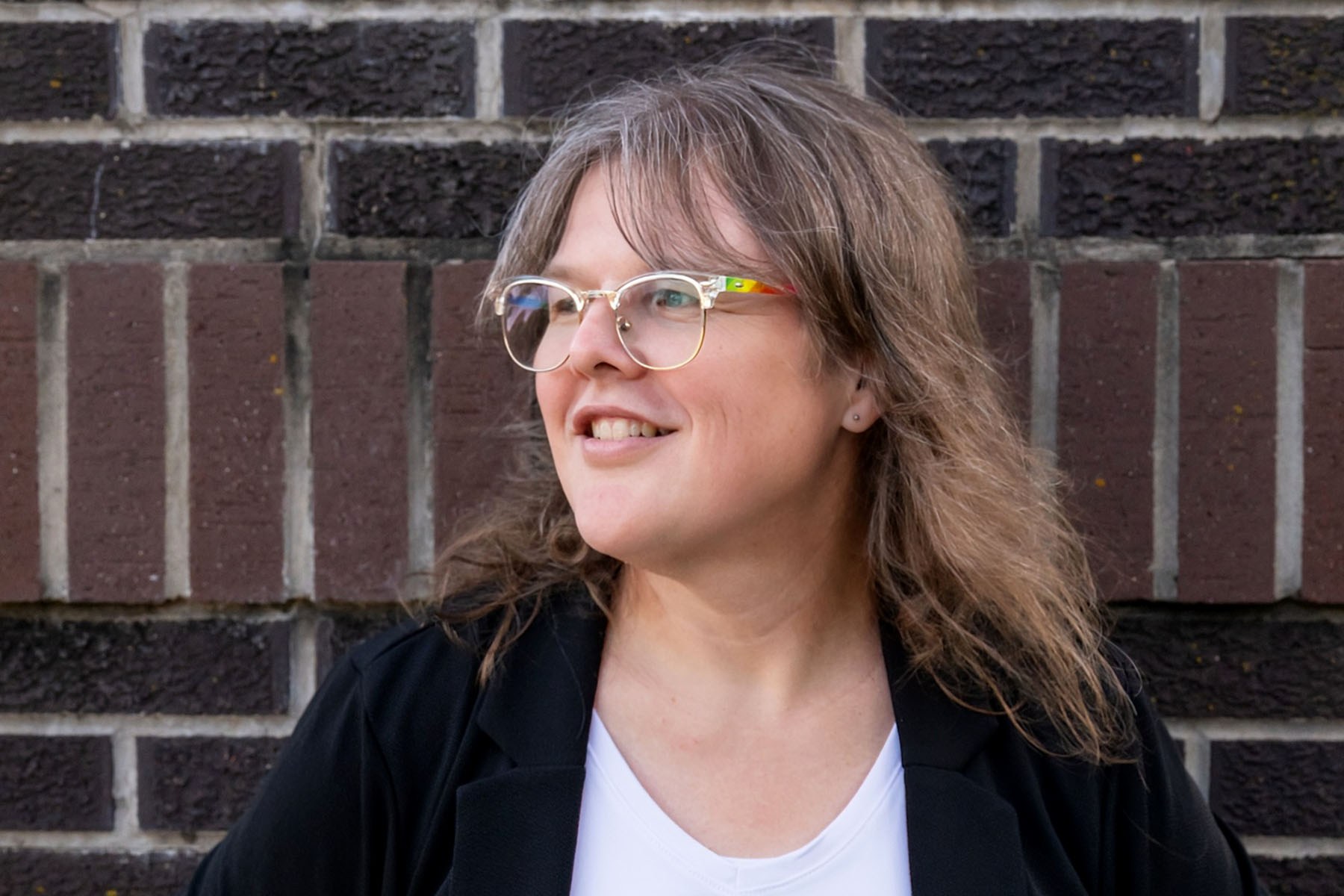 A person with shoulder-length light brown hair and glasses smiles while looking to the left of the frame. They wear a black blazer over a white shirt and stand in front of a dark red and black brick wall.