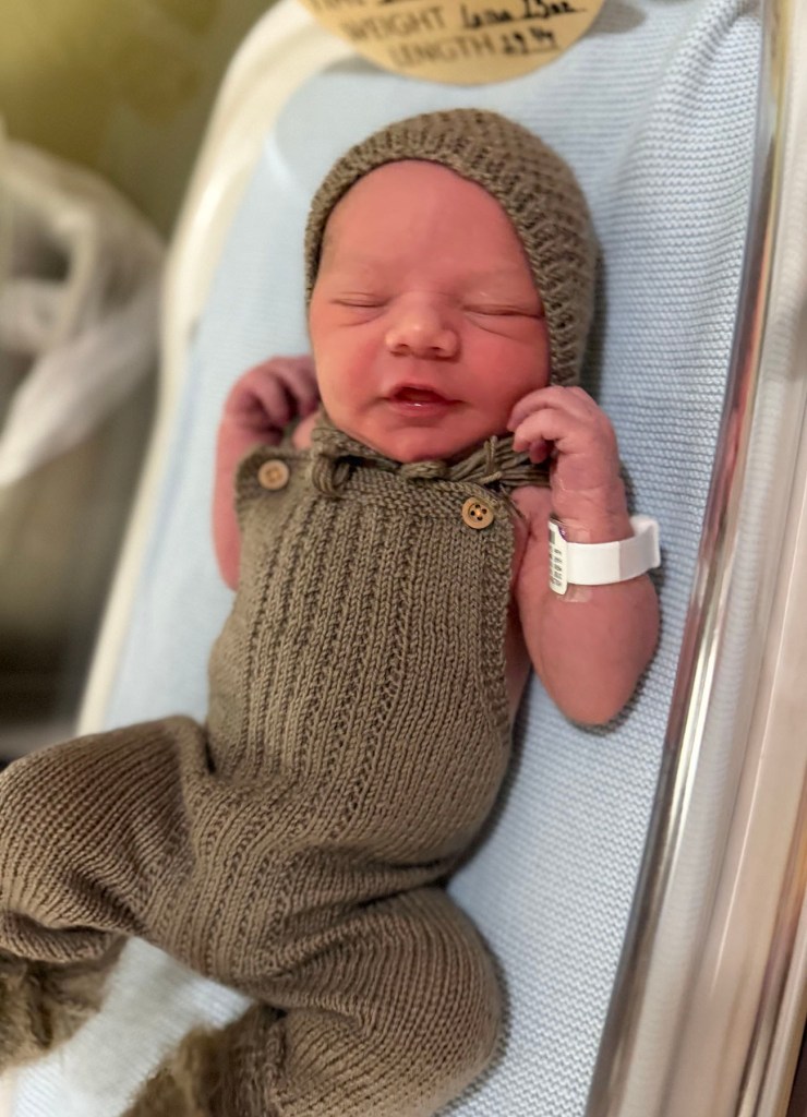 A newborn baby sleeps in a hospital bassinet, wearing a brown knit hat and matching outfit, with a hospital identification band around one wrist.
