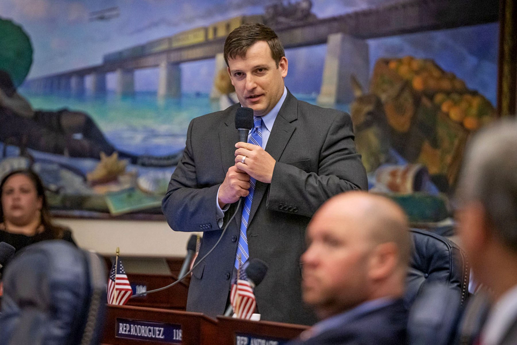 Mike Beltran, wearing a gray suit and blue striped tie, speaks into a microphone in a legislative chamber.