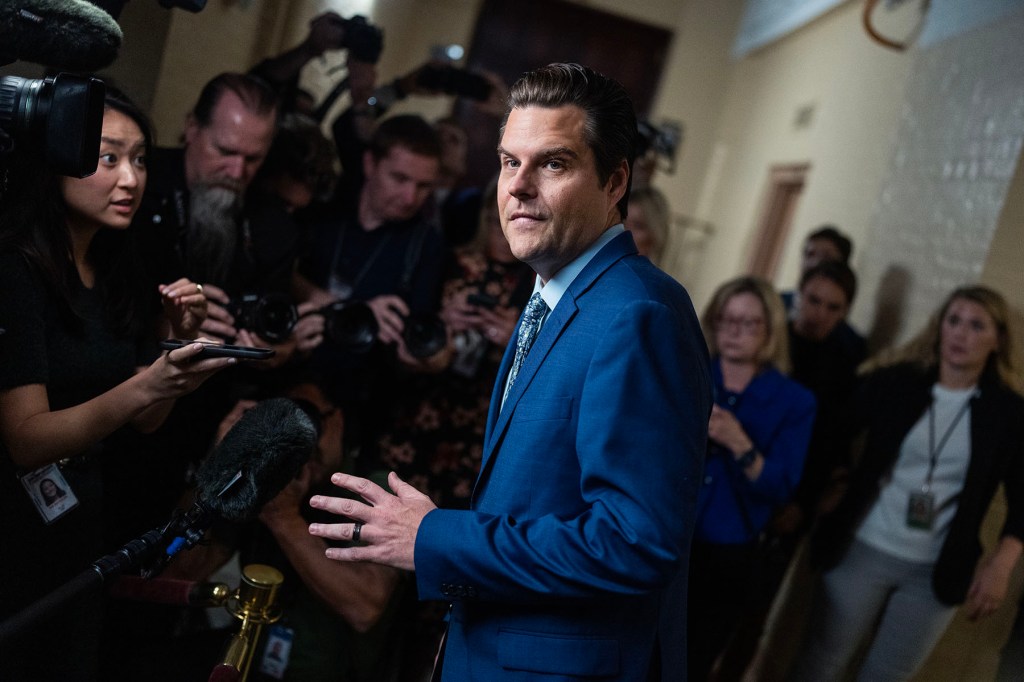 Matt Gaetz, wearing a bright blue suit and patterned tie, faces reporters in a crowded Capitol hallway. Journalists surround him with cameras, microphones and recording devices.