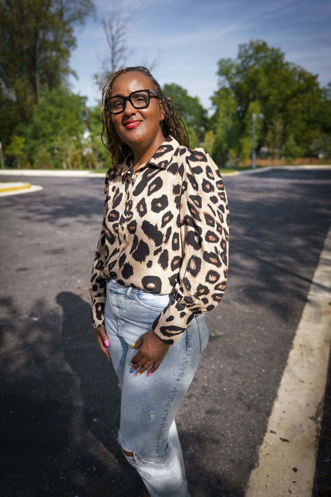 Darnese Foster wearing black-framed glasses, a leopard print blouse and jeans stands outdoors on a sunny residential street.