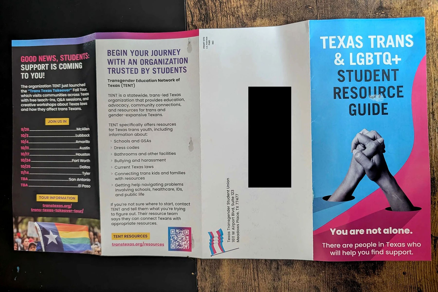An unfolded brochure titled “Texas Trans & LGBTQ+ Student Resource Guide” lies on a wooden surface. The blue and pink cover features an illustration of two clasped hands and the words “You are not alone.”