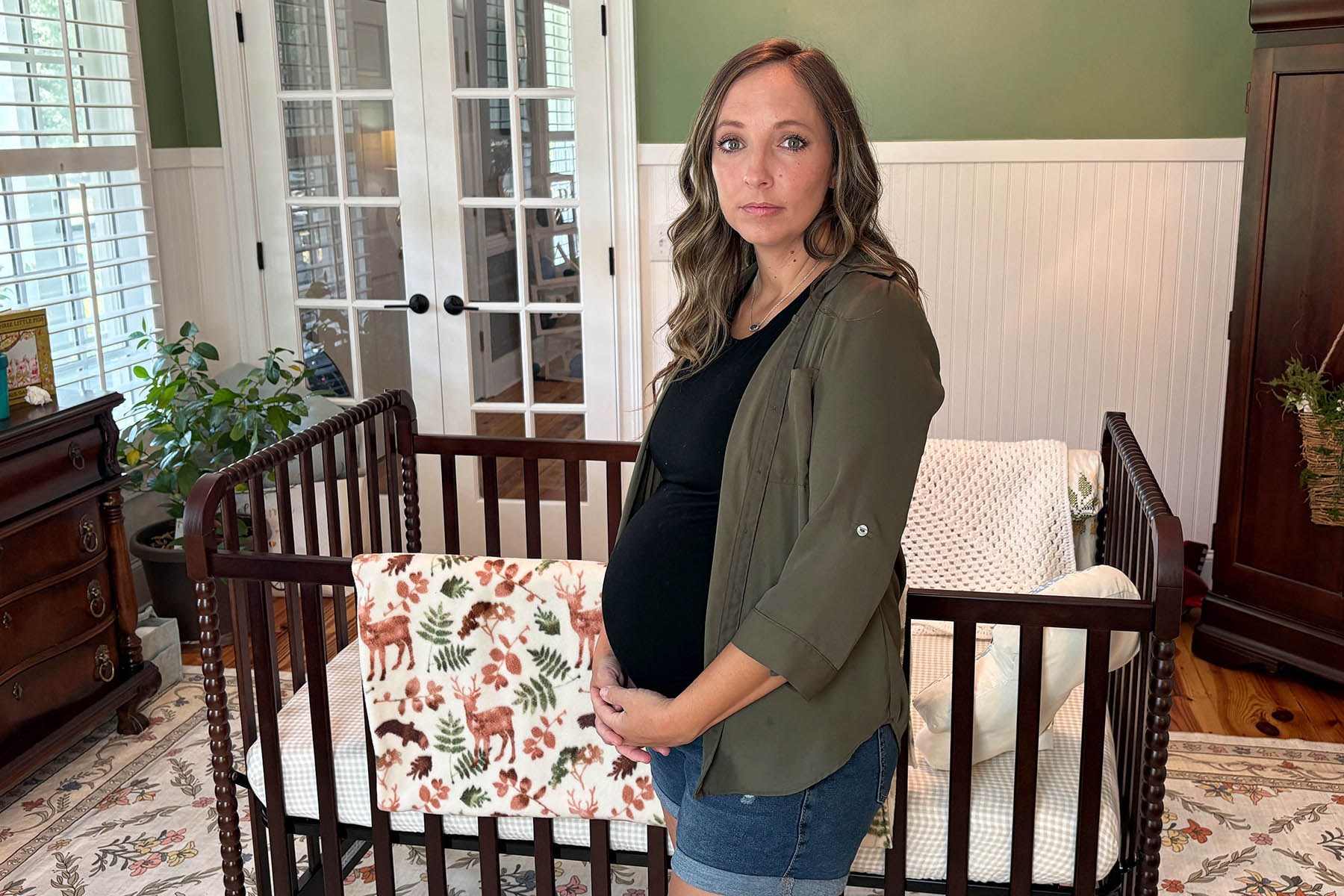 A pregnant woman stands beside a wooden crib in a nursery, resting her hands on her belly and looking toward the camera.
