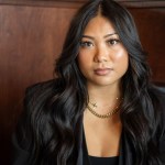A woman with long dark hair wearing a black blazer sits for a portrait against a wood-paneled wall.