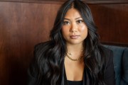 A woman with long dark hair wearing a black blazer sits for a portrait against a wood-paneled wall.