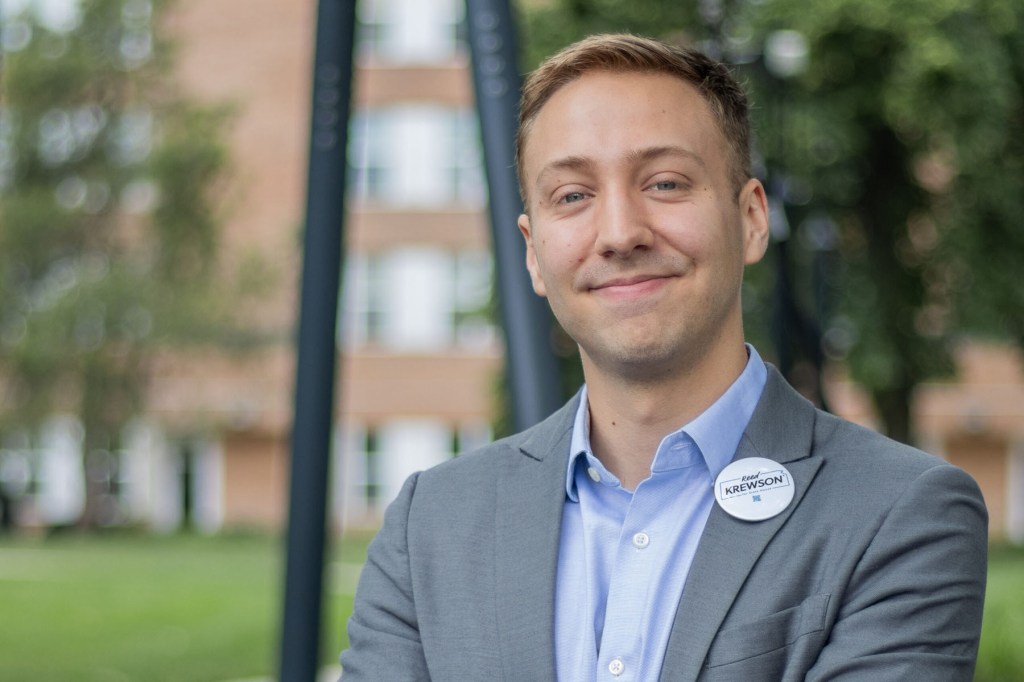 A man in a gray suit and blue shirt smiles for a portrait outdoors, wearing a campaign button that reads “Reed Krewson.”