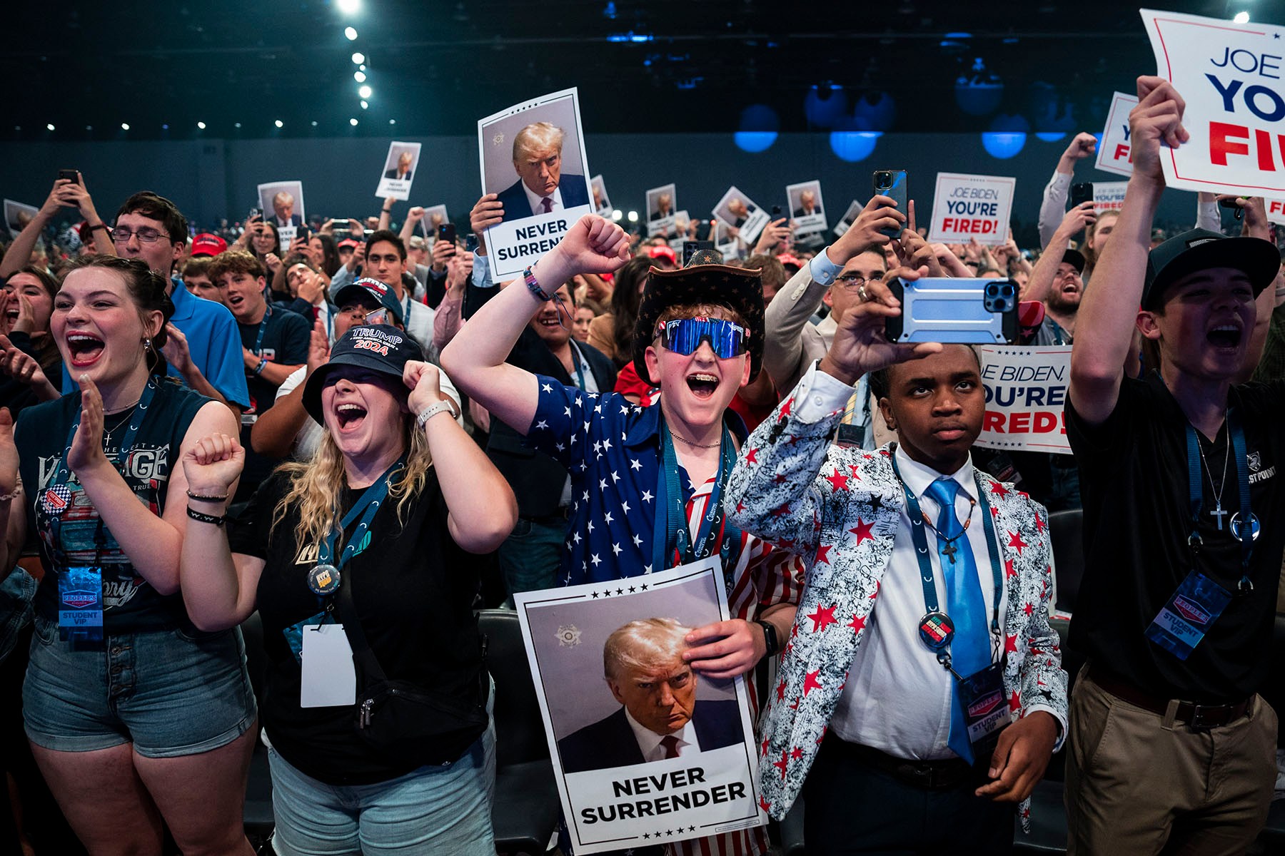 A crowd of cheering young attendees at a Turning Point USA event holds up signs featuring Donald Trump and slogans including "Never Surrender" and "Joe Biden, You're Fired!" as others raise their phones to take photos.