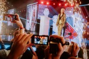 Erika Kirk in a gold sequined suit stands onstage surrounded by pyrotechnics and large video screens as audience members hold up their phones to photograph her.