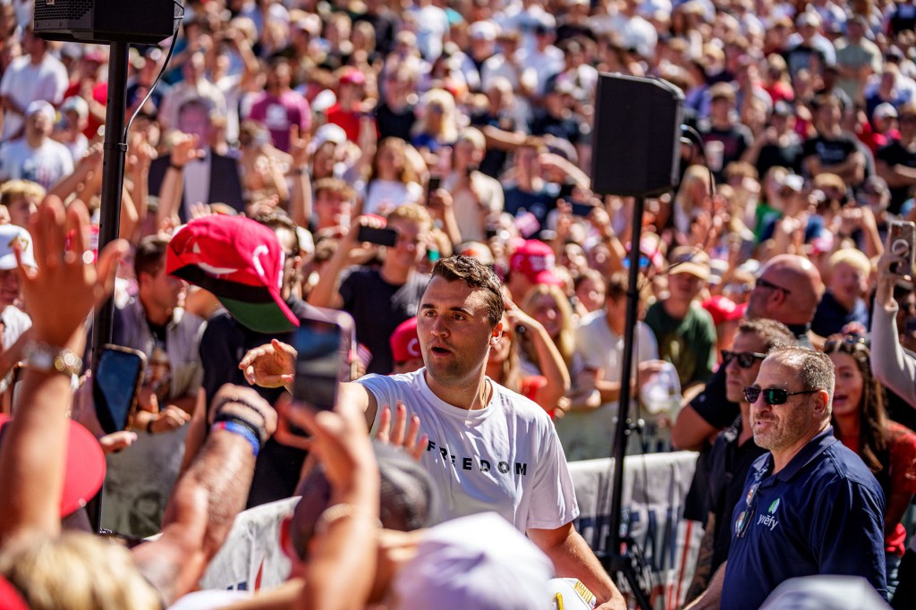Charlie Kirk tosses a red baseball cap toward a crowd of supporters, many reaching toward him or holding up their phones, at an outdoor event.