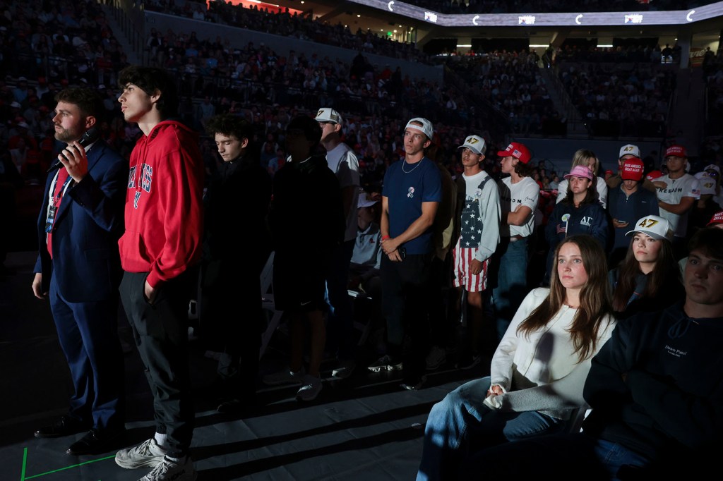 Young attendees wearing Turning Point USA merchandise stand and sit in a dimly lit arena, illuminated by stage lighting.