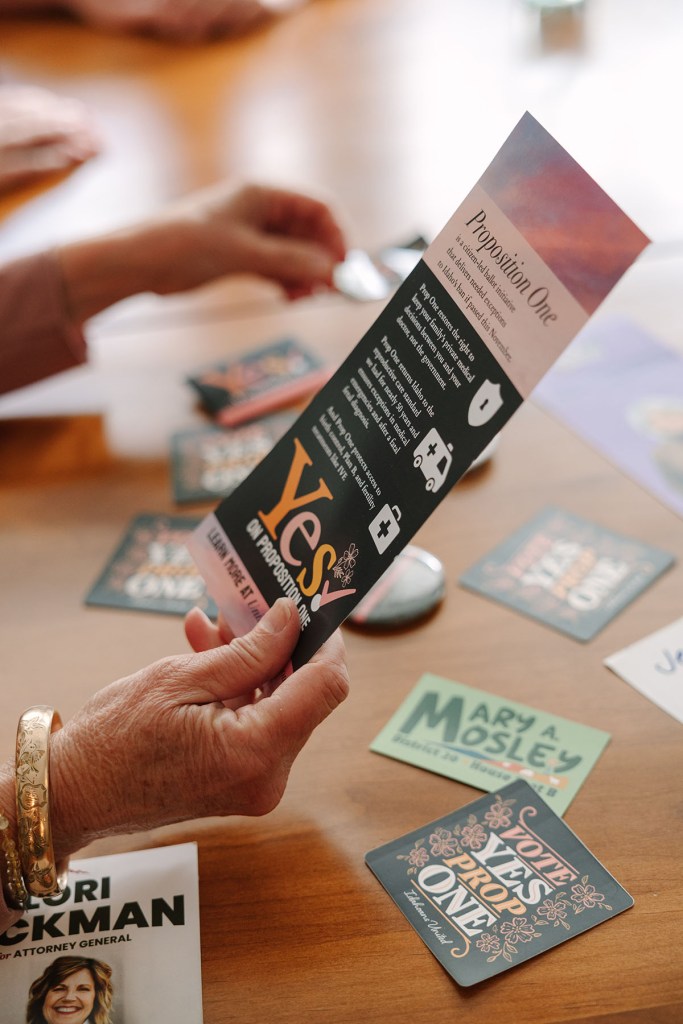 Lisa holds a Proposition One campaign flyer above a table scattered with political stickers and campaign materials.