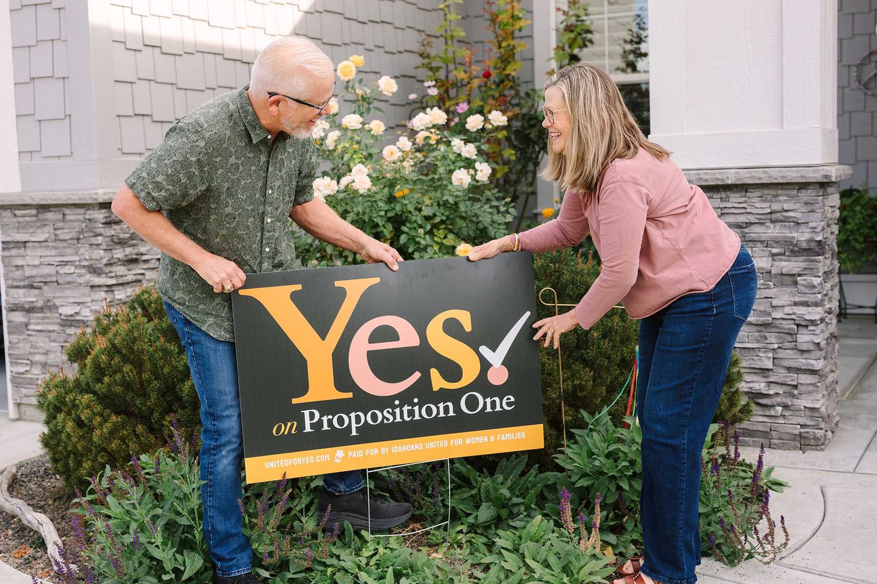 Steve and Lisa smile at each other as they place a “Yes on Proposition One” sign in their front garden.