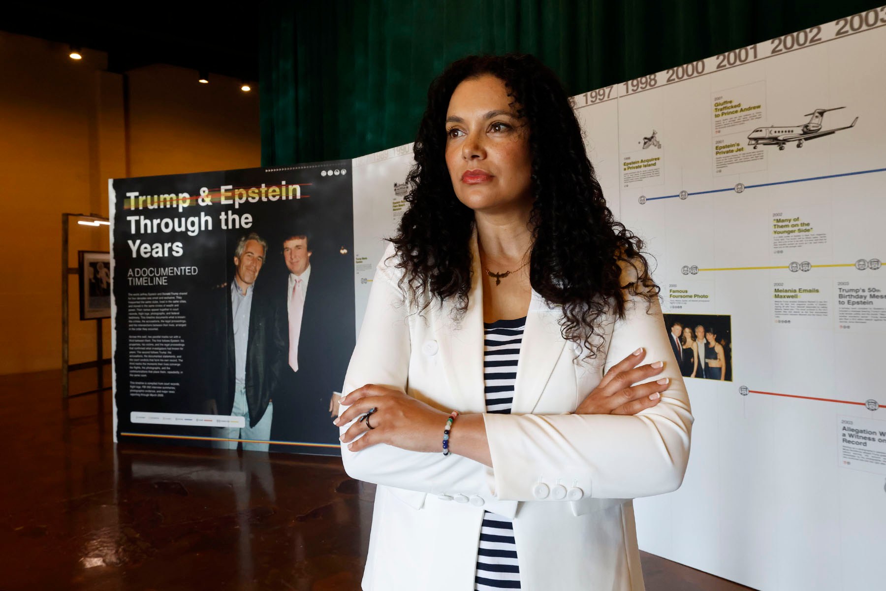 Sharlene Rochard stands with her arms crossed in front of a timeline documenting Donald Trump's relationship with Jeffrey Epstein at an exhibition in Dallas. She wears a white blazer over a striped shirt.