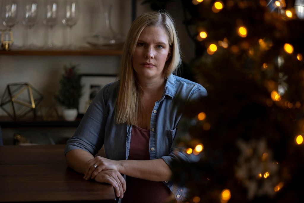 Kaitlyn Kash sits at a wooden table in her home, wearing a denim shirt over a pink top.
