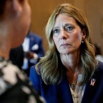 Amy Acton, wearing a navy blazer with an Ohio flag pin, listens intently to a person in the foreground.