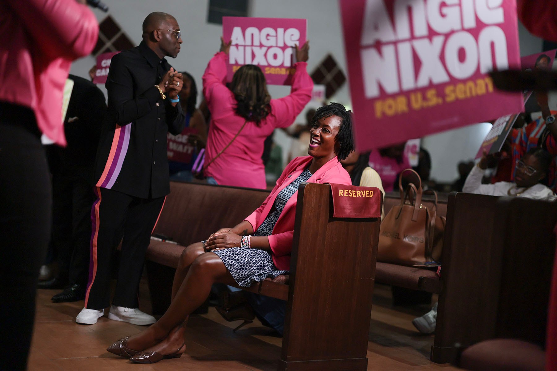 Angie Nixon laughs while seated in a church pew at a campaign event, wearing a bright pink blazer. Supporters around her hold pink Angie Nixon for U.S. Senate signs.