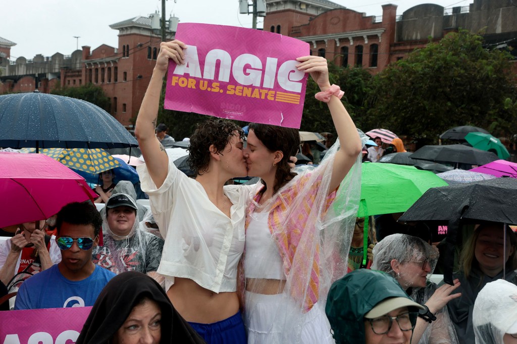 Two supporters kiss beneath an Angie Nixon for U.S. Senate sign at an outdoor rally in the rain. A crowd holding colorful umbrellas surrounds them, with several attendees wearing ponchos.