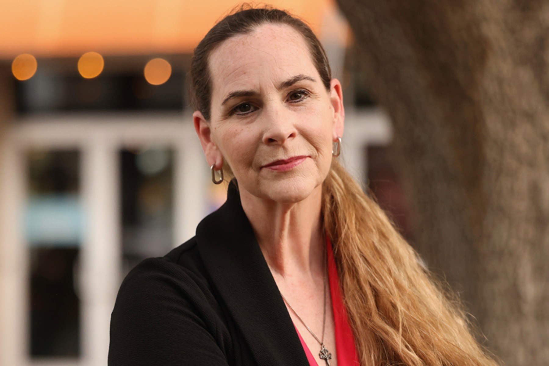 A woman with long, light brown hair pulled back into a ponytail looks directly at the camera. She wears a black blazer over a red top, silver hoop earrings and a necklace.