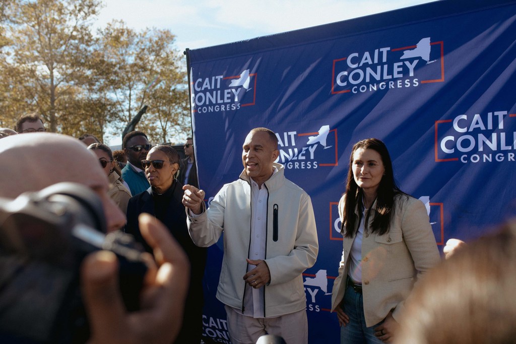 House Minority Leader Hakeem Jeffries gestures toward a crowd while standing beside Cait Conley in front of a blue campaign backdrop. Photographers and supporters gather around them.