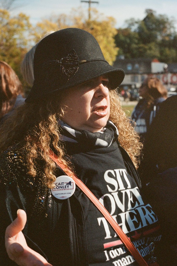 Mellicent Dyane, wearing a black cloche hat, patterned scarf and Cait Conley campaign button, speaks outdoors at a rally. Her shirt reads, "Sit over there. I don't need your mansplaining."