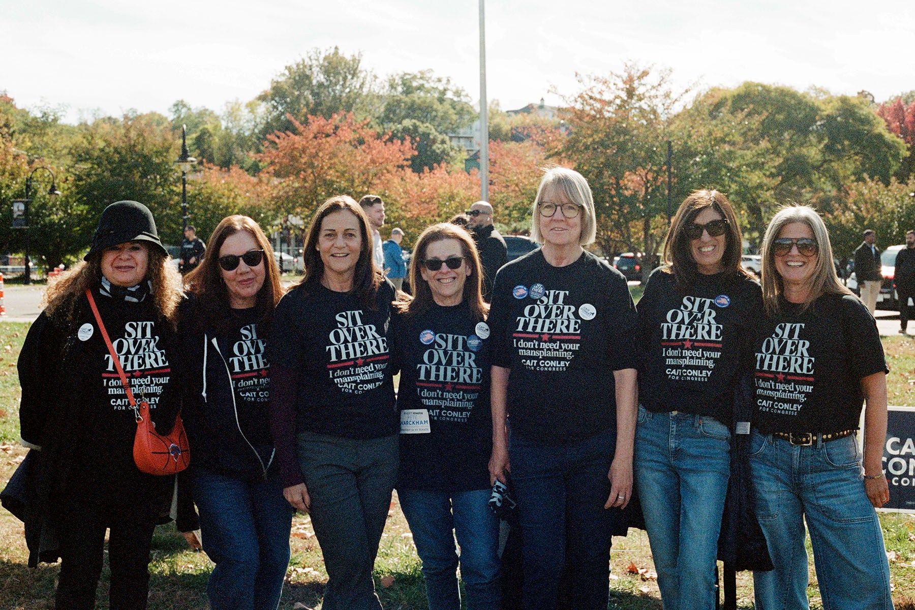 Six women stand together outdoors wearing black Cait Conley campaign shirts that read, "Sit over there. I don't need your mansplaining." Autumn trees and other rallygoers are visible behind them.