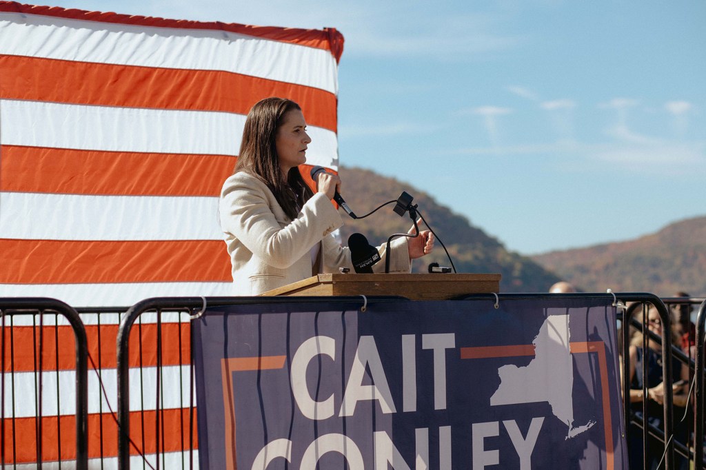 Cait Conley speaks into a handheld microphone behind a campaign podium, with a large American flag hanging behind her and wooded hills in the distance.