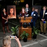 A woman speaks at a podium crowded with microphones during a nighttime press conference, surrounded by several people holding notebooks.