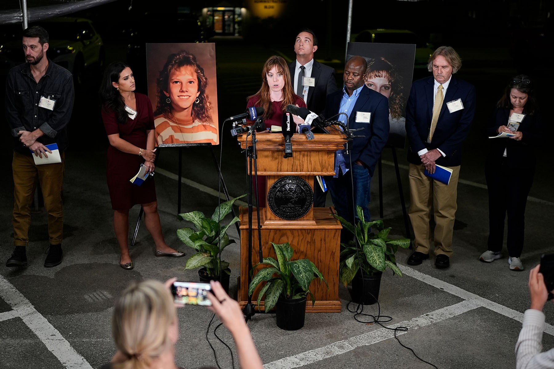 A woman speaks at a podium crowded with microphones during a nighttime press conference, surrounded by several people holding notebooks.