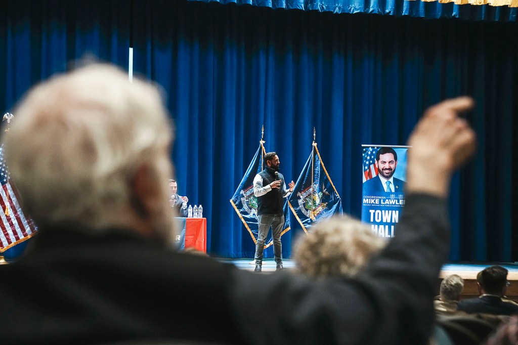 Republican Rep. Mike Lawler speaks into a microphone on a stage framed by blue curtains and New York state flags. An audience member raises a hand in the foreground, partially obscuring the stage.