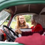 Rebecca Cooke leans into the open window of a vintage vehicle to speak with an older couple seated inside.