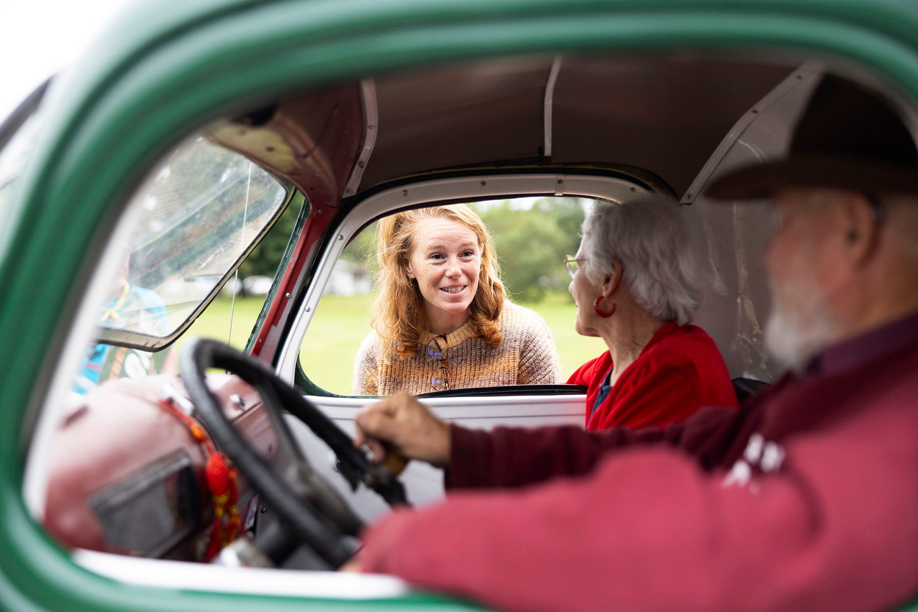 Rebecca Cooke leans into the open window of a vintage vehicle to speak with an older couple seated inside.