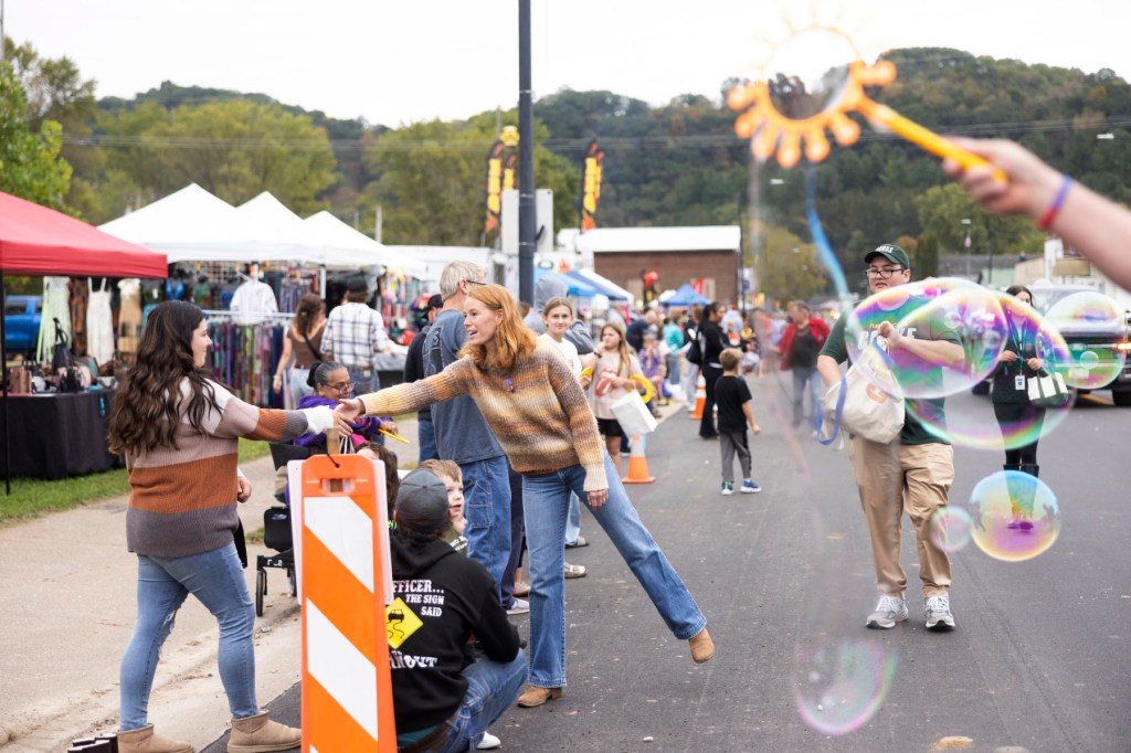 Rebecca Cooke reaches out to greet a festivalgoer on a crowded street as large bubbles float nearby.