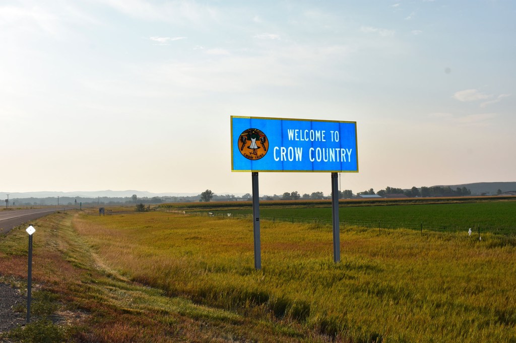 A blue roadside sign reading "Welcome to Crow Country" stands beside a road cutting through open grassland.