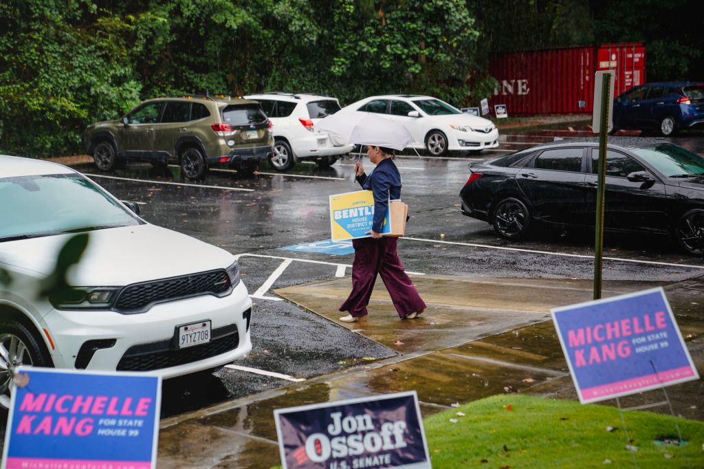 Hudgins walks through a parking lot in the rain with an umbrella, surrounded by campaign materials.