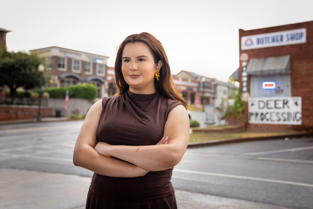 Portrait of Bautista standing arms crossed in the middle of an empty rural street with a building behind her that says "Deer processing"