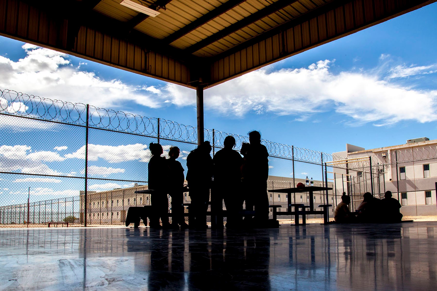 Silhouettes of people gather around picnic tables beneath a covered outdoor area at a detention facility. Chain-link fences topped with razor wire and large buildings surround the space.
