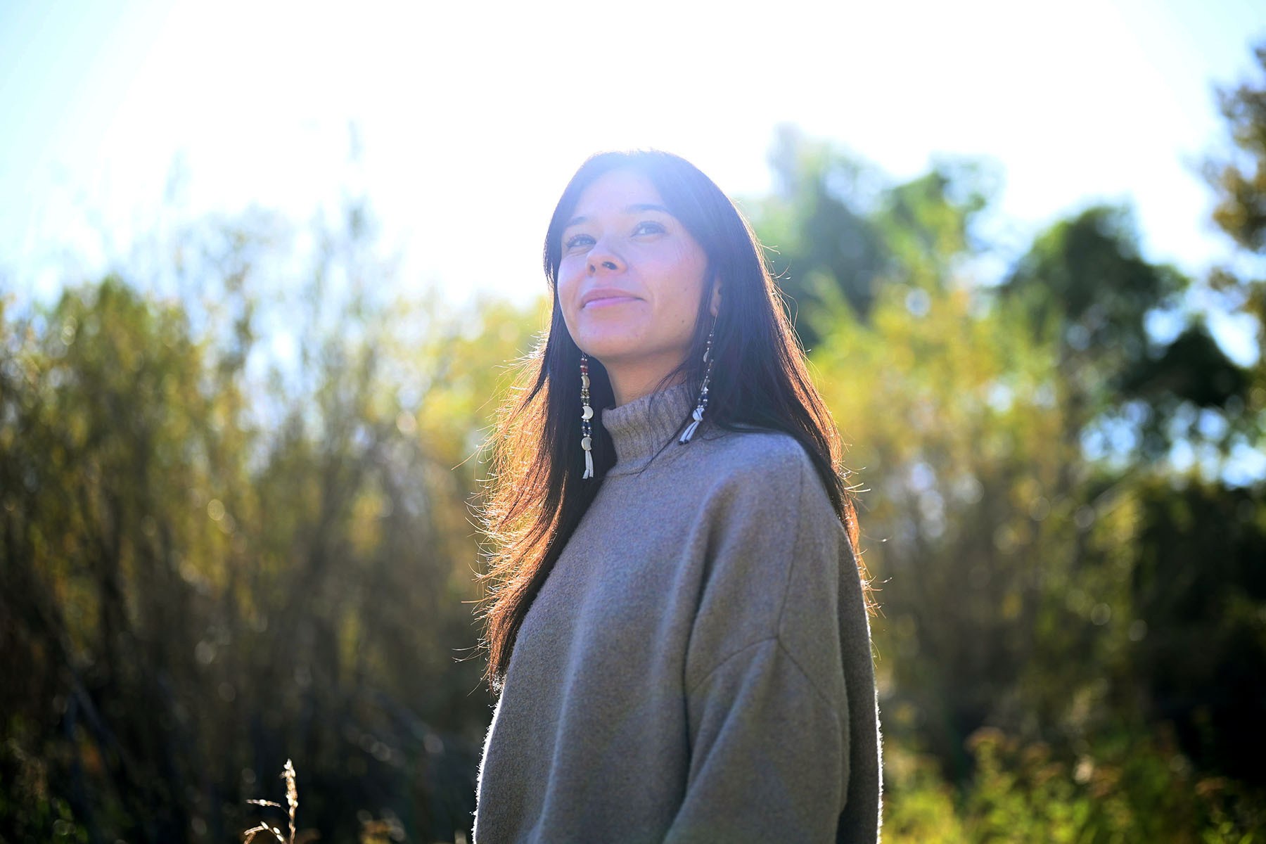 Katie Fire Thunder stands outdoors in a gray sweater, looking upward and slightly to the right. Sunlight filters through the trees behind her, illuminating her hair.