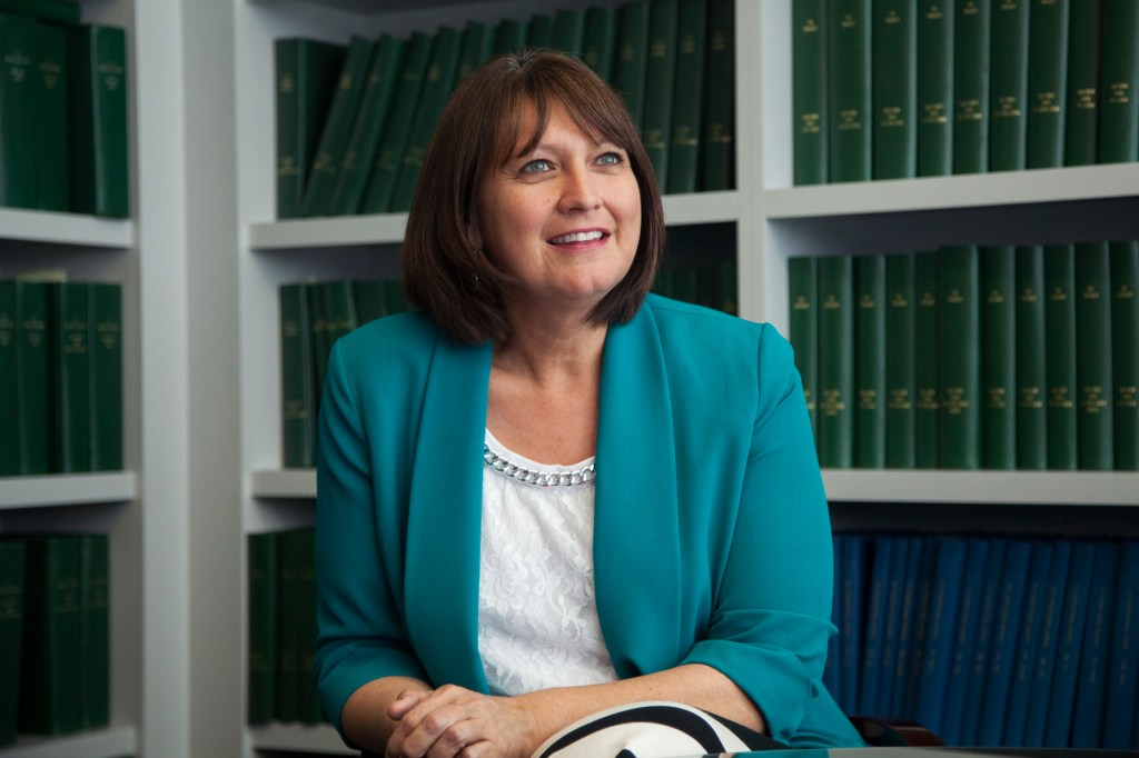 Denise Juneau sits smiling in front of shelves filled with green books, wearing a turquoise blazer over a white blouse.