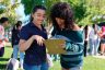 A person helps another person fill out a form outside in a grassy area surrounded by other people.