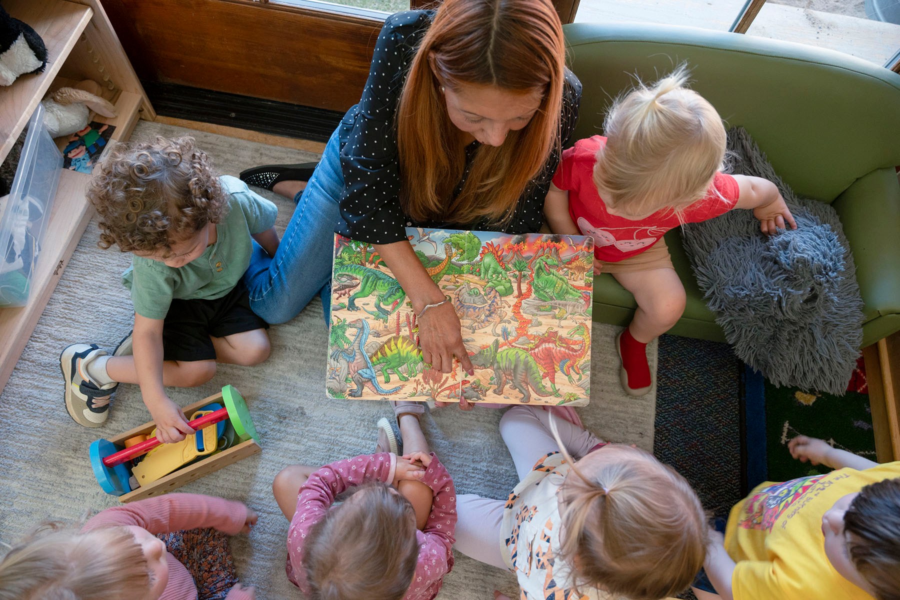 Overhead view of a woman reading a colorful dinosaur book to several young children gathered around her in a childcare setting.