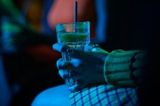 Close-up of a person wearing rings holding a clear drink with a lime wedge and straw in a dimly lit bar, illuminated by blue and green light.