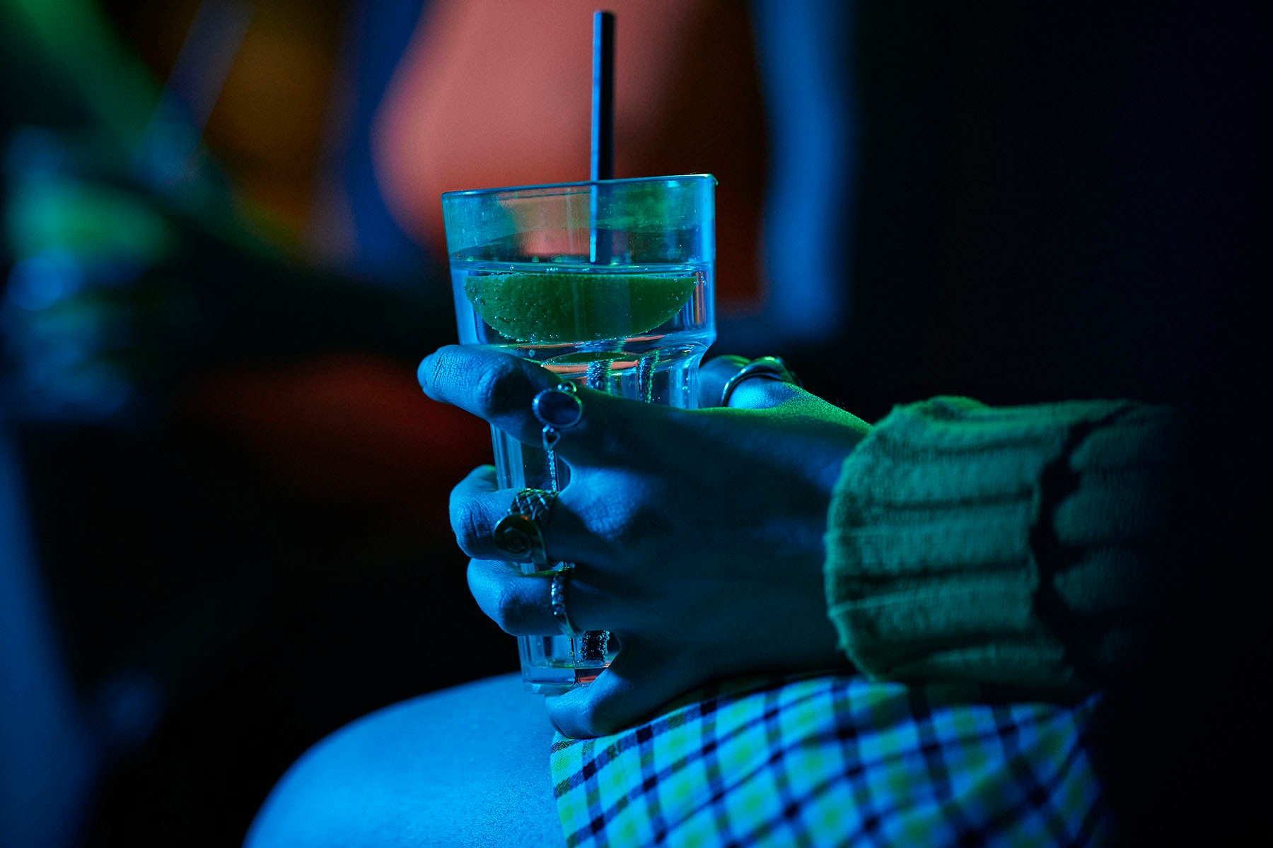Close-up of a person wearing rings holding a clear drink with a lime wedge and straw in a dimly lit bar, illuminated by blue and green light.