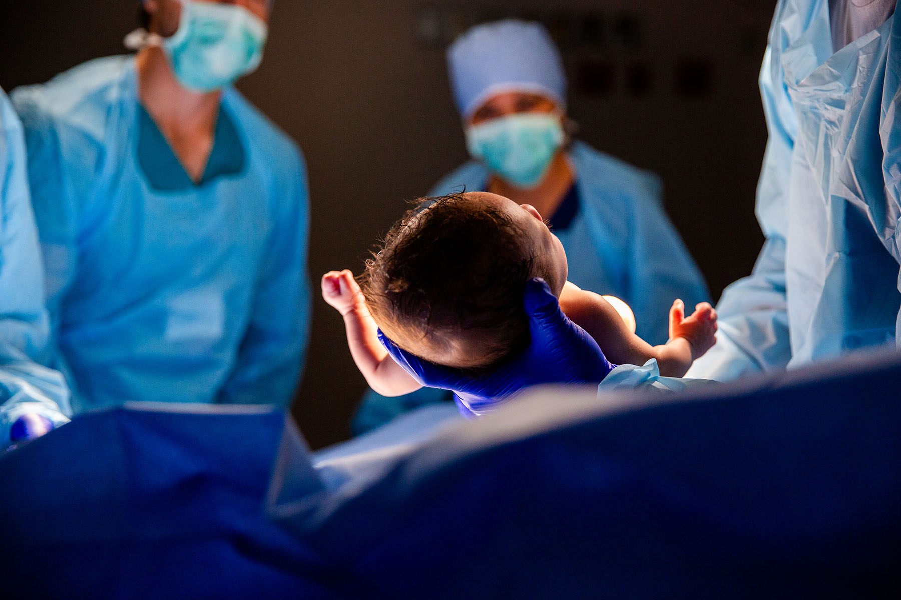 A newborn is held up by a gloved medical worker in an operating room as masked staff look on in the background.