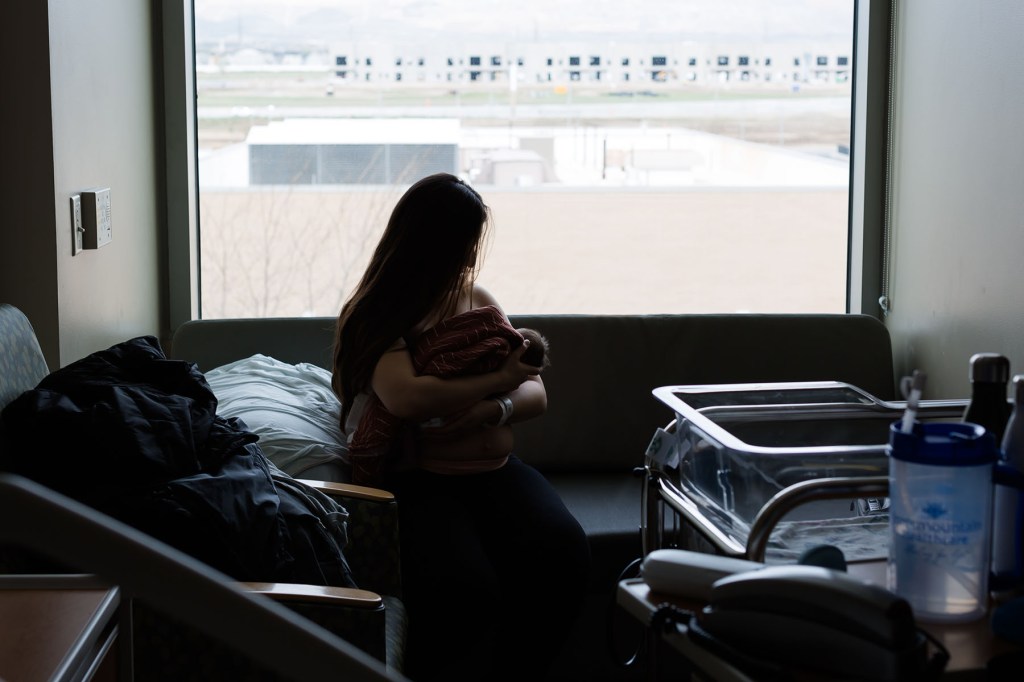 A person sits in a hospital room holding a newborn against their chest, silhouetted in front of a large window.