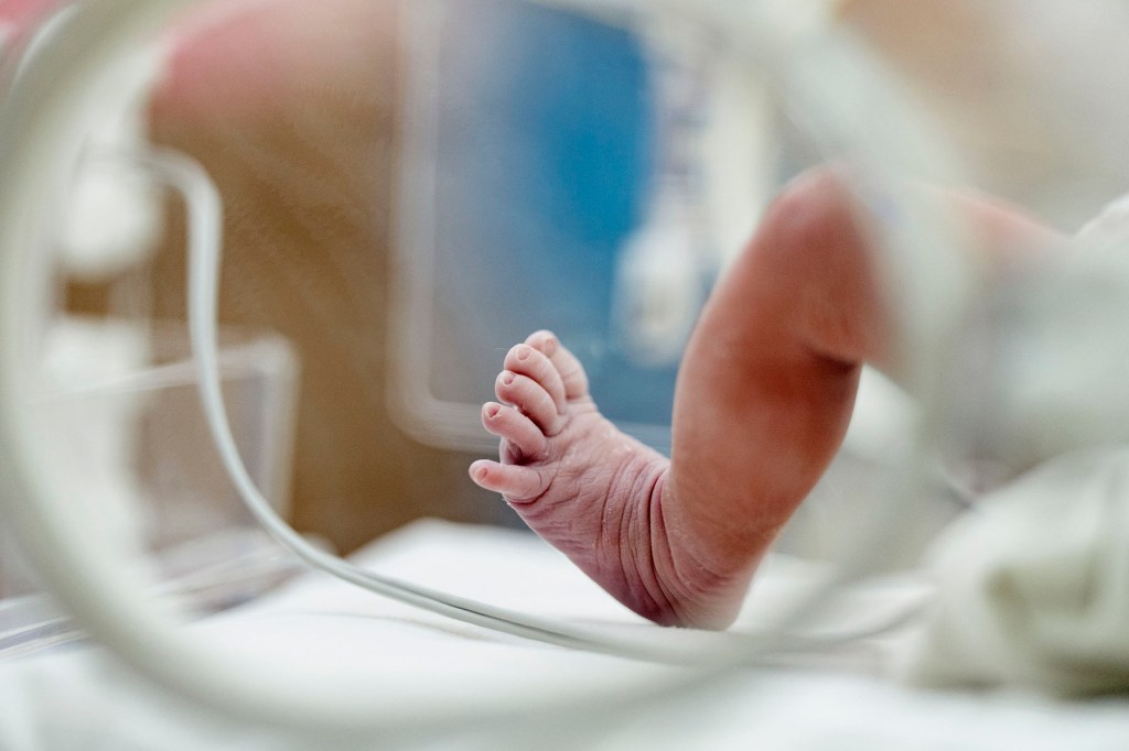Close-up of a newborn’s foot resting inside an incubator, with medical equipment blurred in the background.