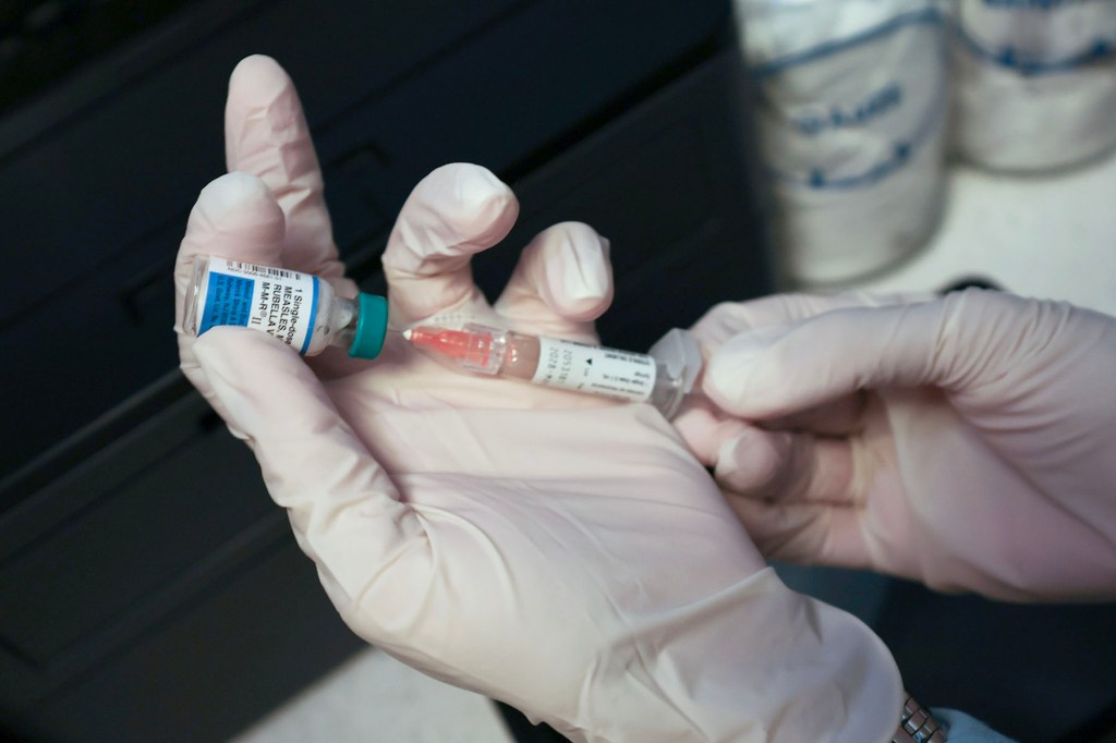 Gloved hands draw a measles vaccine dose into a syringe from a vial.