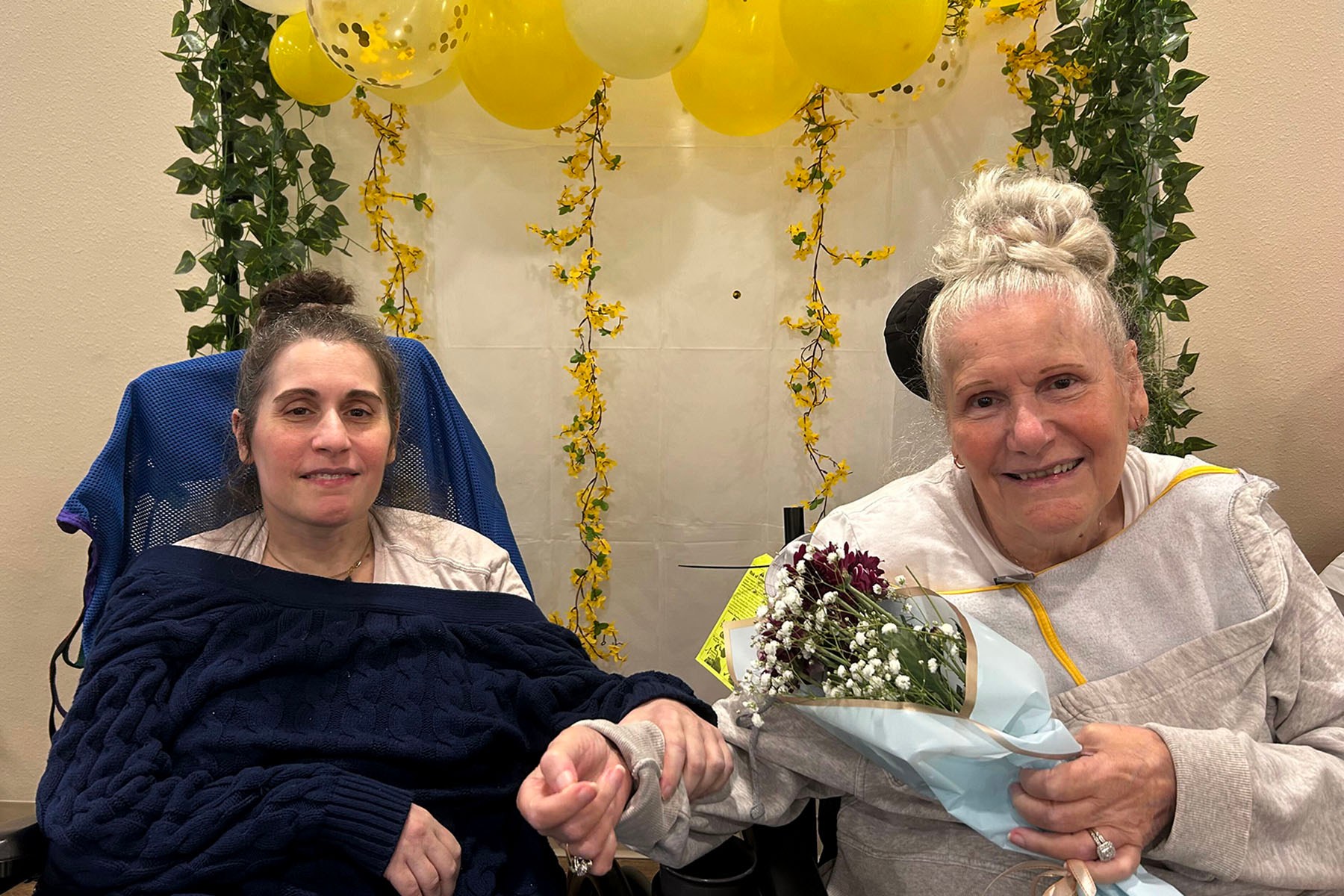 Melissa Napolitan (left) and her mother, Cindy, sit side by side holding hands. Cindy holds a bouquet of flowers, and yellow balloons and hanging greenery decorate the wall behind them.